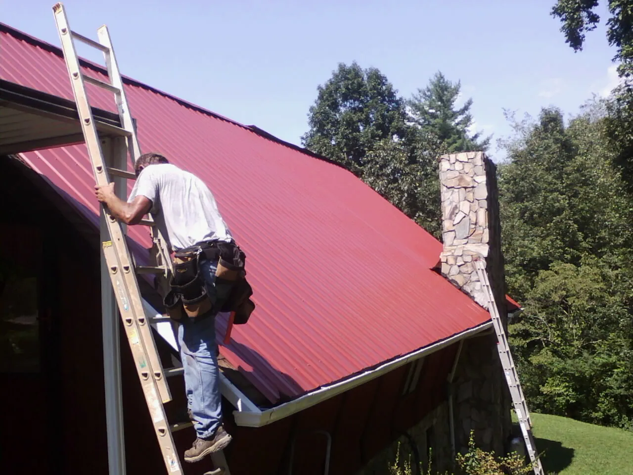 Roofer on a ladder beside a red metal roof and stone chimney — Soffit & Fascia Repair by Roofing Brothers in Pearson